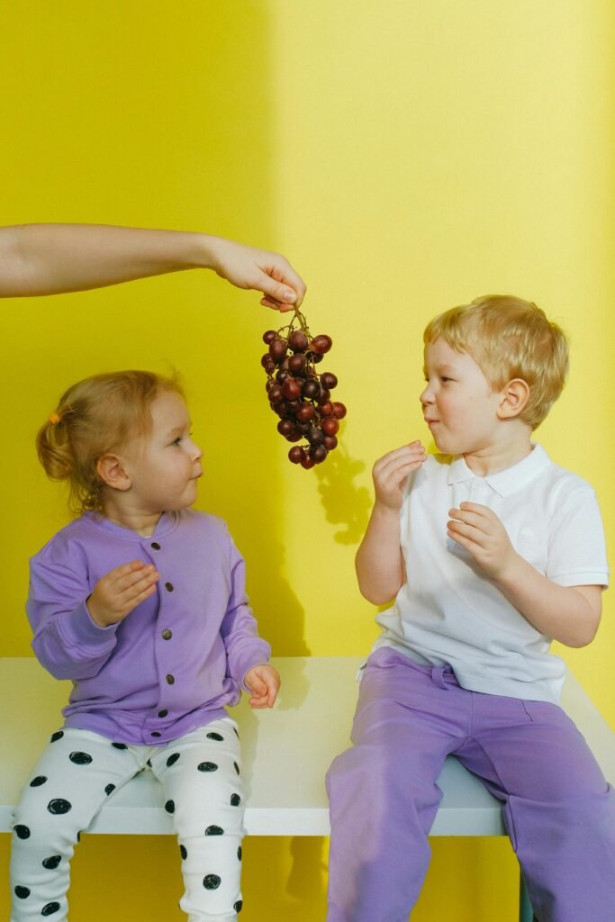 Two young children sitting indoors enjoying grapes against a bright yellow background.